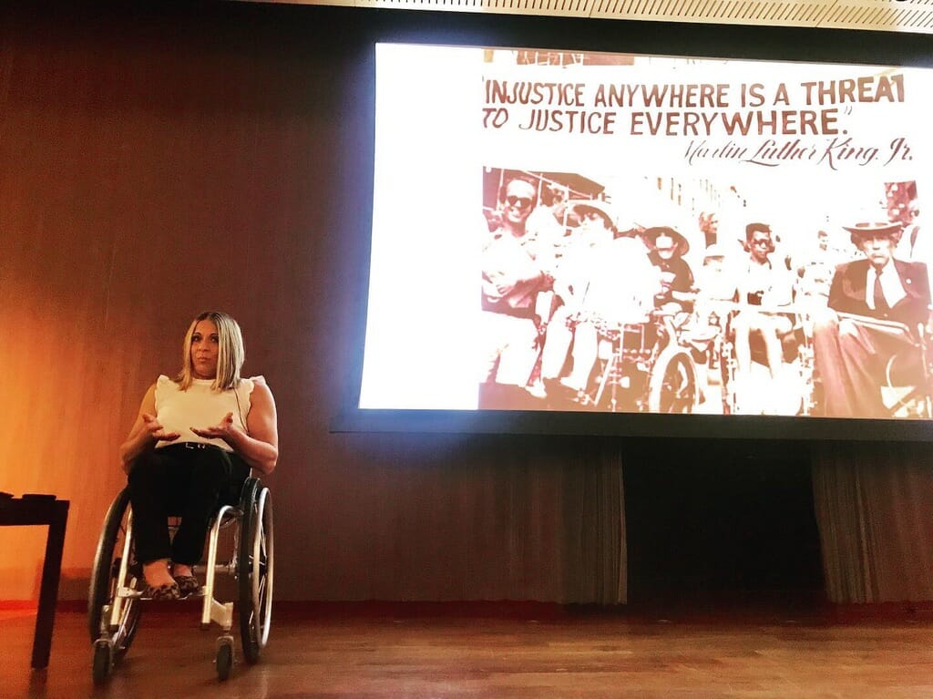 alycia delivering speech at asu on stage with large screen displaying disability history image